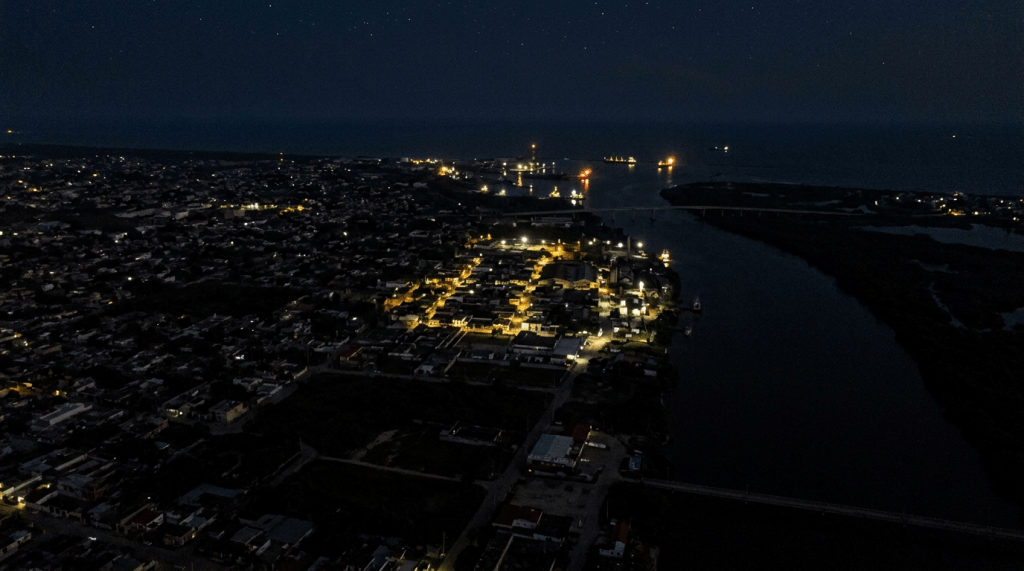 A nighttime aerial view of a coastal city with illuminated streets and buildings near a harbor and river