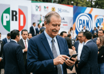 Man in suit checking smartphone at political event with PRI and PAN banners behind