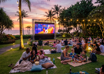 Outdoor movie screening of Coco with families sitting on blankets and chairs on grass near ocean