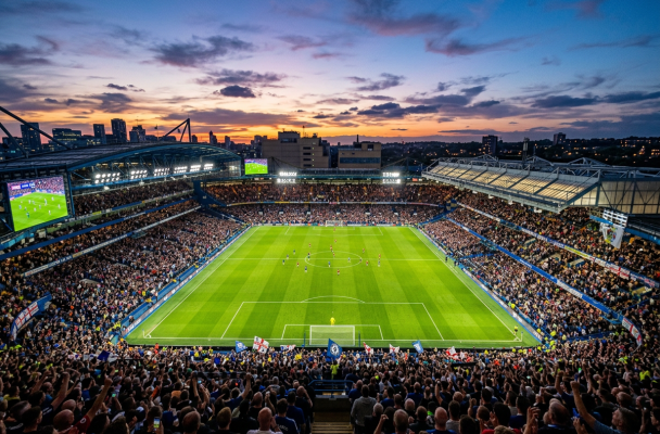 A packed stadium during a football match at sunset with players on the field and cheering fans.