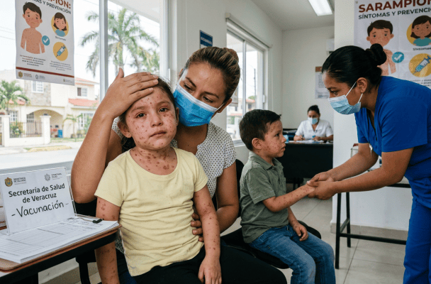 Children receiving care at a vaccination clinic; posters read “Sarampión: síntomas y prevención” and “Vacunación.”